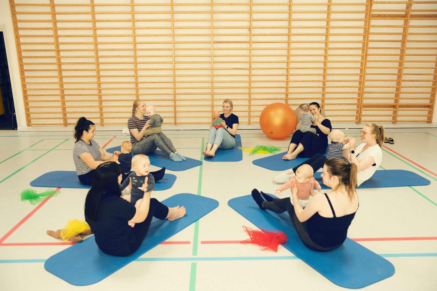 Mødre og børn samlet i en gymnastiksal sammen med Ergoterapeuten Monica. De laver øvelser med en stor bold.