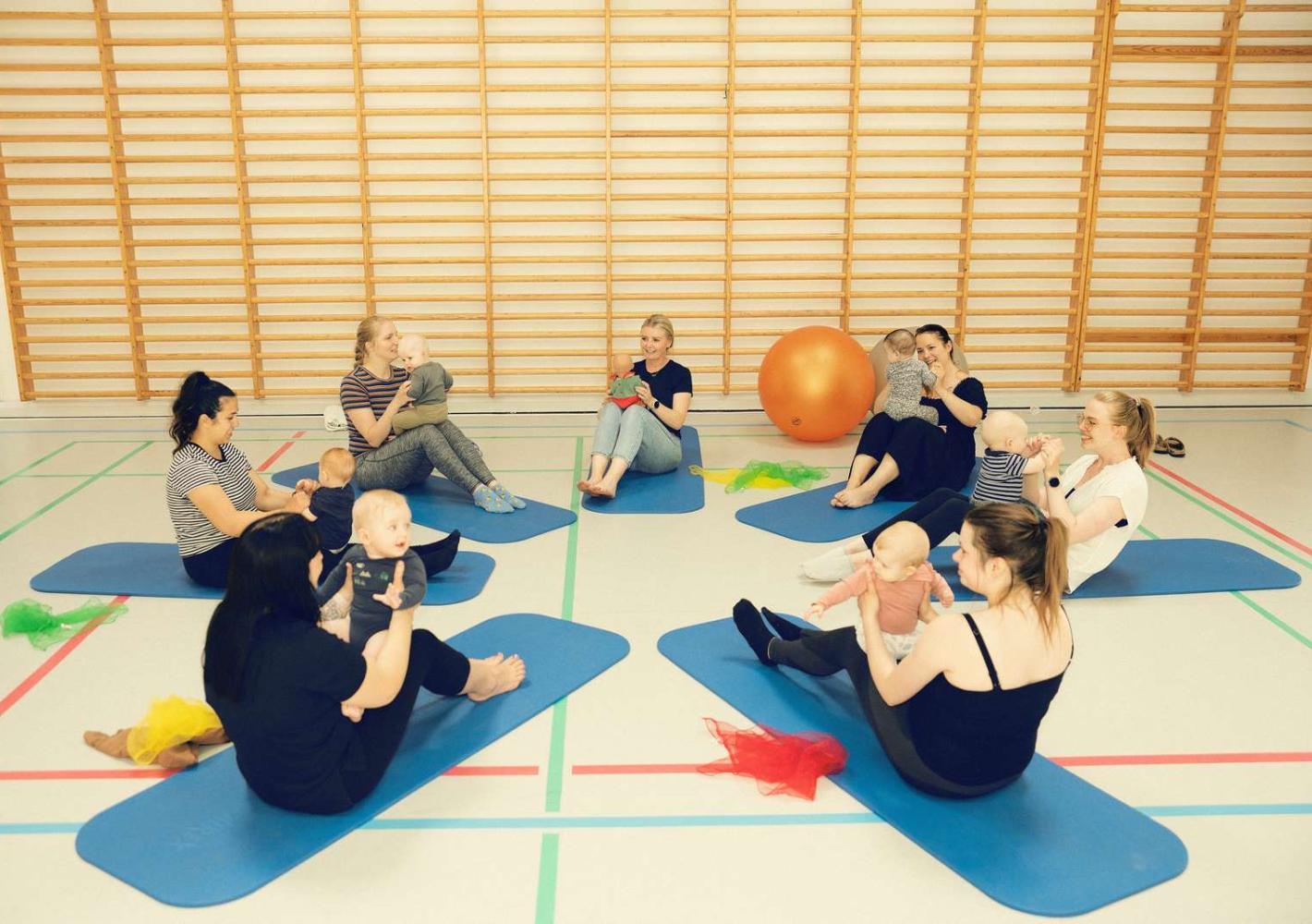 Mødre og børn samlet i en gymnastiksal sammen med Ergoterapeuten Monica. De laver øvelser med en stor bold.