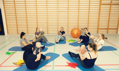 Mødre og børn samlet i en gymnastiksal sammen med Ergoterapeuten Monica. De laver øvelser med en stor bold.